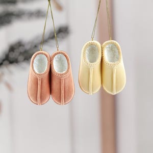 Könnte beinhalten: Zwei Paar dekorative Pantoffel-Ornamente, eines in Pfirsich und eines in Hellgelb. Beide haben ein flauschiges weißes Innenfutter und sind an goldfarbenen Schnüren aufgehängt, ideal für die Weihnachtsdekoration.