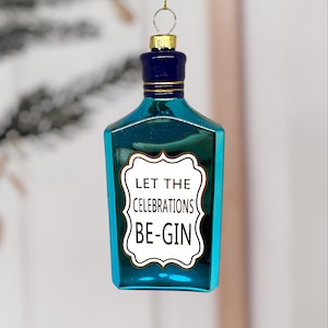 Può includere: Ornamento natalizio in vetro color verde acqua a forma di bottiglia di gin. L'ornamento ha un tappo blu scuro con dettagli dorati e un'etichetta bianca con la scritta "LET THE CELEBRATIONS BE-GIN". Un anello dorato in alto per appenderlo.
