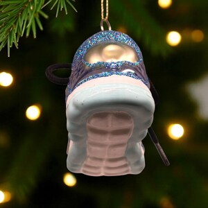 May include: A Christmas tree ornament shaped like a sneaker. The ornament is primarily light blue and white, with dark blue accents and glitter. A black shoelace and loop are visible. The ornament hangs from a gold string.