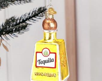 Può includere: Un ornamento di vetro a forma di bottiglia di tequila. La bottiglia è gialla con un tappo dorato e un'etichetta che recita "Tequila 100% Agave".