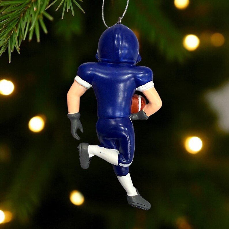 Peut inclure: Ornement repr&eacute;sentant un joueur de football am&eacute;ricain bleu, tenant un ballon marron et blanc, en pleine course. L'ornement a des d&eacute;tails blancs sur les manches et les chaussettes, avec des gants et des chaussures gris. L'arri&egrave;re-plan est un sapin de No&euml;l flou.