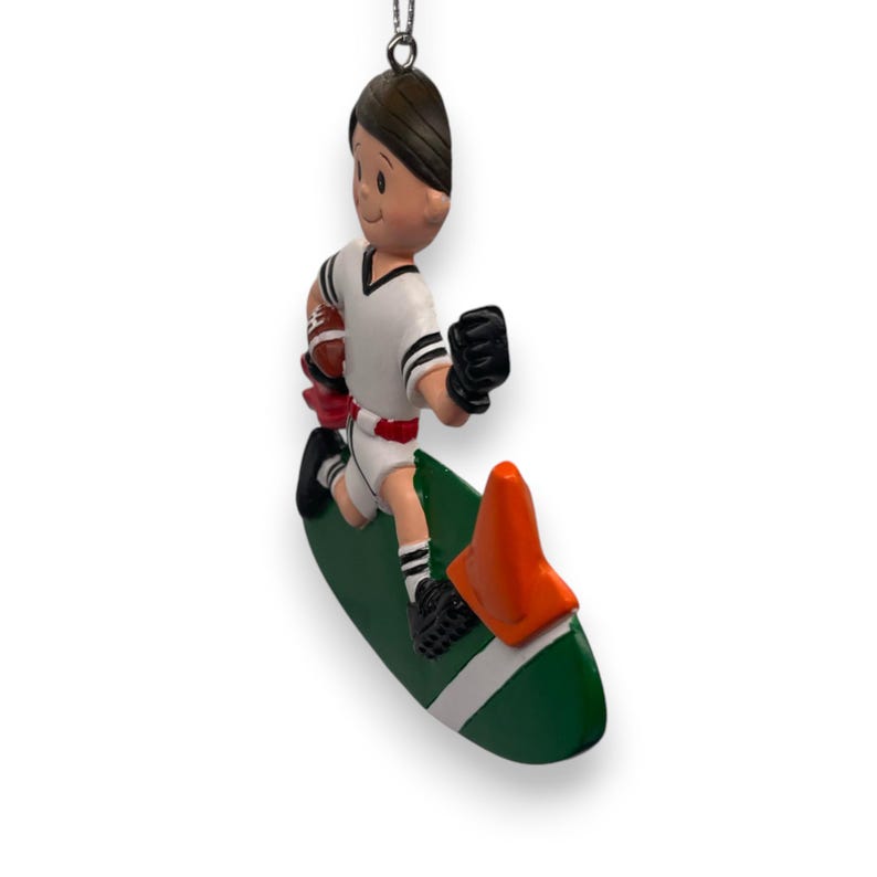 May include: A green and white football field ornament with a cartoon football player running with a ball. The player is wearing a white jersey with black stripes and black pants. The ornament is topped with an orange traffic cone.
