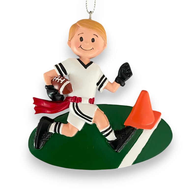 May include: A personalized Christmas ornament featuring a cartoon boy wearing a white and black football uniform, holding a football and running with a red flag attached to his waist. The boy is running towards an orange cone on a green field with a white line.