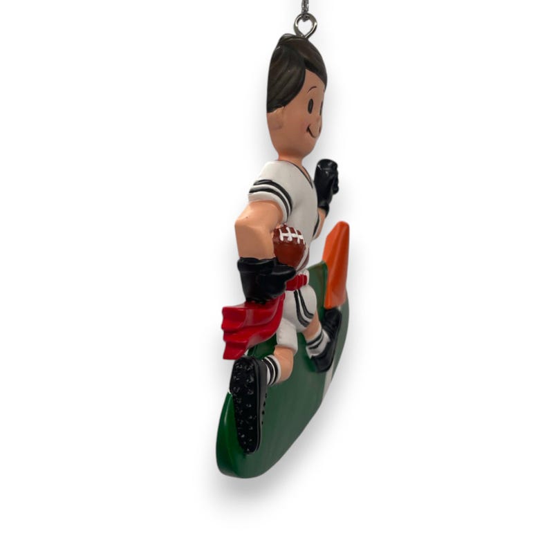 May include: A white and green Christmas ornament depicting a football player running with a football in hand. The player is wearing a white jersey with black stripes, black pants, and a red and white scarf. The ornament is shaped like a football field.