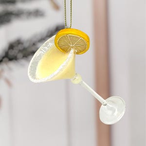 May include: A clear glass margarita ornament with a yellow drink and a sugared rim. A yellow lemon slice is attached to the rim. The ornament hangs from a gold string.