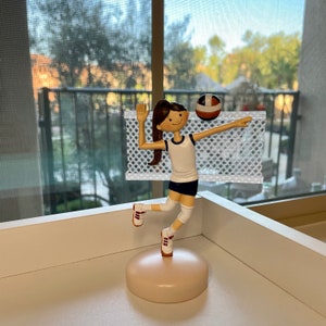 Puede incluir: Una figura de una jugadora de voleibol con cabello casta&ntilde;o, vistiendo una camiseta blanca con ribetes azules, pantalones cortos blancos y zapatillas blancas. La jugadora est&aacute; sosteniendo un bal&oacute;n de voleibol y est&aacute; saltando frente a una red blanca.
