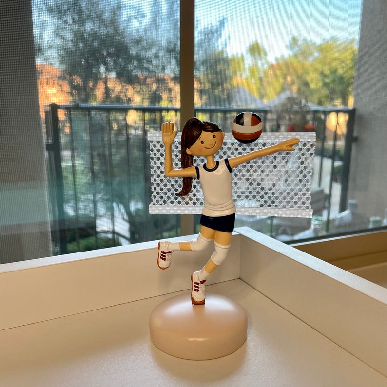 Puede incluir: Una figura de una jugadora de voleibol con cabello casta&ntilde;o, vistiendo una camiseta blanca con ribetes azules, pantalones cortos blancos y zapatillas blancas. La jugadora est&aacute; sosteniendo un bal&oacute;n de voleibol y est&aacute; saltando frente a una red blanca.