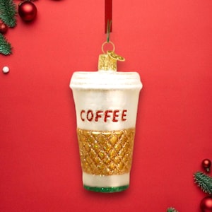 May include: A white glass ornament shaped like a coffee cup with a gold glitter design and the word "COFFEE" in red lettering.