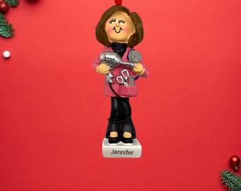 Op de afbeelding: Een gepersonaliseerde kerstversiering met een vrouw met bruin haar die een föhn en een schaar vasthoudt. De versiering is roze en zilverkleurig met de naam "Jennifer" op de basis. De versiering is gedateerd "2025".