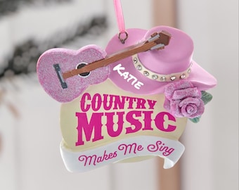 May include: A pink country music-themed ornament featuring a guitar, a cowboy hat, and a rose. The ornament has the text "COUNTRY MUSIC Makes Me Sing" and the name "KATIE". The guitar and hat are embellished with glitter.