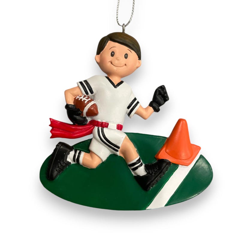 May include: A personalized Christmas ornament featuring a cartoon boy running with a football on a green field. He is wearing a white jersey with black stripes, black shorts, and a red ribbon. There is an orange traffic cone on the field.