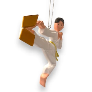 May include: A white karate ornament with a yellow belt, kicking a gold-colored block. The ornament is designed to hang on a Christmas tree.