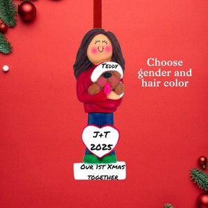 May include: A personalized Christmas ornament featuring a figure holding a teddy bear wearing a Santa hat. The ornament has dark hair, a red sweater, blue pants, and a heart with initials and the year 2025. The base reads "OUR 1ST XMAS TOGETHER."