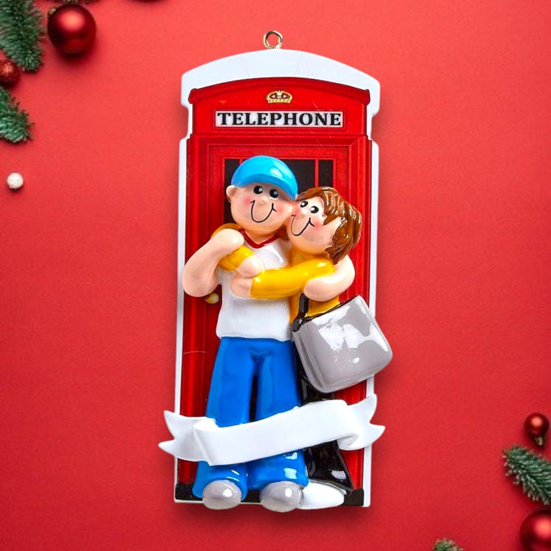 May include: A red telephone box Christmas ornament with a white banner. A couple are standing in front of the box, hugging. The man is wearing a blue hat and the woman is wearing a yellow jumper.