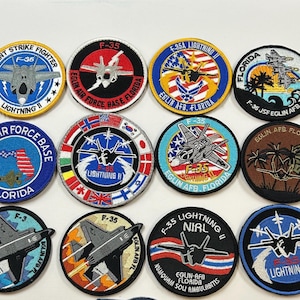 May include: A collection of embroidered patches featuring the F-35 Lightning II fighter jet. The patches are circular, with various designs including the aircraft, text, and emblems. Colors include blue, red, yellow, and black.