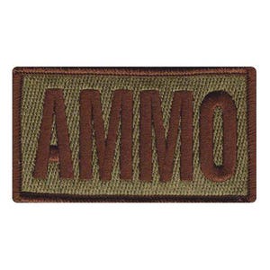 May include: A rectangular embroidered patch with the word "AMMO" in brown thread. The text is stitched onto a khaki-colored fabric, framed by a brown border.
