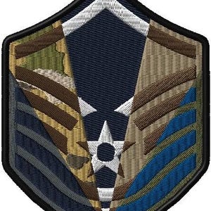 Puede incluir: Un parche bordado en azul, marrón y verde con una estrella de cinco puntas en el centro. El parche tiene forma de escudo y un borde negro.