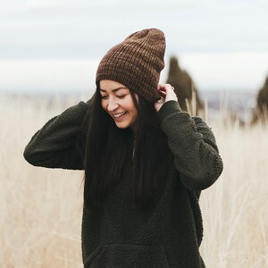 Op de afbeelding: Een vrouw draagt ​​een bruine gebreide beanie en een donkergroene fleece trui met een rits. Ze staat in een veld met hoog, droog gras.
