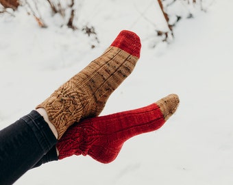 Peut inclure: Une paire de chaussettes tricotées, l'une brune avec un motif géométrique et l'autre rouge avec un motif en côtes. Les chaussettes sont portées par une personne assise dans la neige.