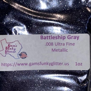 May include: A close-up of a bag of Battleship Gray .008 Ultra Fine Metallic glitter. The bag is labeled with the name of the glitter, the size, and the website address: https://www.gamfunkyglitter.us. The glitter is a dark gray color with a metallic sheen.