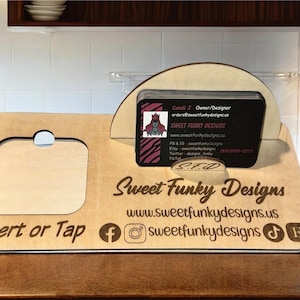 May include: A wooden phone stand with a cutout for a phone and a business card holder. The stand is engraved with the text "Sweet Funky Designs" and the website address "www.sweetfunkydesigns.us". The stand also features social media icons for Facebook, Instagram, TikTok, and Etsy.