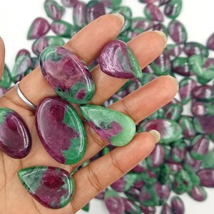 May include: A collection of green and pink gemstone cabochons in a teardrop shape. The stones are arranged in a pile and a hand is holding a few of the stones.