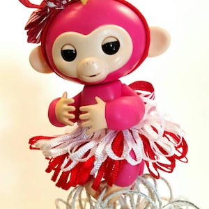 May include: A pink monkey figurine with a red and white striped bow and a red and white ribbon skirt. The monkey has a light-coloured face and ears, and is standing on a decorative silver base.