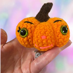 May include: A small, orange crocheted pumpkin ornament featuring a dark brown stem, large green plastic eyes with black pupils, embroidered black eyelashes, and a bright pink stitched mouth and nose detail.