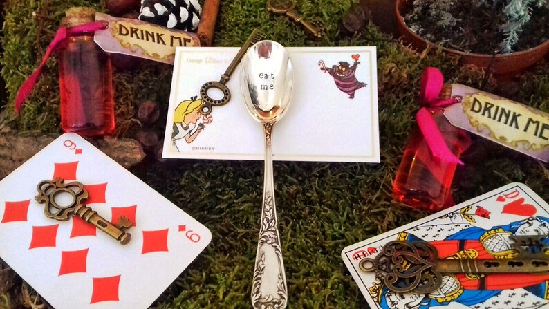 Pu&ograve; includere: Una scena a tema Alice nel Paese delle Meraviglie con un cucchiaio d'argento con la scritta "eat me", carte da gioco, chiavi e piccole bottiglie con etichette "drink me".