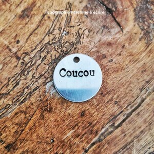 Pu&ograve; includere: Un piccolo tag rotondo in metallo argentato con la parola "Coucou" stampata in grassetto. Il tag ha un piccolo foro nella parte superiore per l'attacco. Lo sfondo &egrave; una superficie di legno. Il testo "Typographie Machine &agrave; &eacute;crire" &egrave; anche visibile.