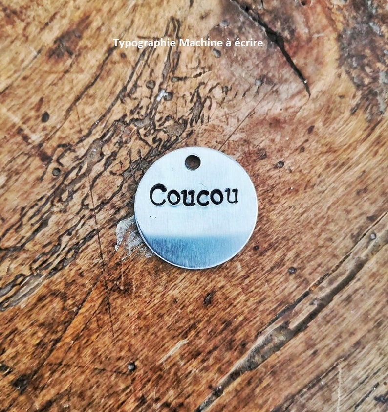 Pu&ograve; includere: Un piccolo tag rotondo in metallo argentato con la parola "Coucou" stampata in grassetto. Il tag ha un piccolo foro nella parte superiore per l'attacco. Lo sfondo &egrave; una superficie di legno. Il testo "Typographie Machine &agrave; &eacute;crire" &egrave; anche visibile.