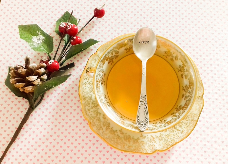 Pu&ograve; includere: Una tazza e un piattino vintage con bordi dorati, riempiti di t&egrave;, e un cucchiaino d'argento con la parola "love" stampata. Un rametto di bacche e una pigna sono sul lato, su una superficie rosa a forma di cuore.