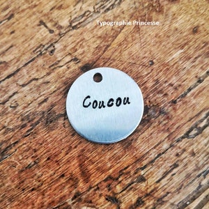 Pu&ograve; includere: Un piccolo ciondolo rotondo in metallo color argento con un foro nella parte superiore. La parola "Coucou" &egrave; scritta in corsivo nero al centro del ciondolo. Il ciondolo poggia su una superficie di legno.
