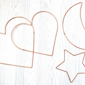 May include: Four copper wire shapes: a crescent moon, a star, and two overlapping hearts. The shapes are on a white wooden background.