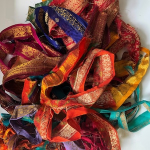 May include: A collection of assorted fabric ribbons featuring vibrant colours including red, orange, yellow, purple, blue, and teal. Each ribbon displays intricate gold metallic patterns and decorative borders with frayed edges throughout the pile.