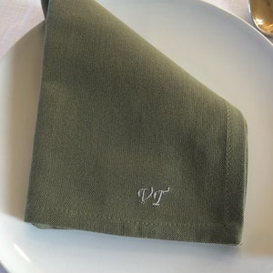 Peut inclure: Une serviette en lin verte pliée en triangle avec les initiales "V.T." brodées en fil blanc.