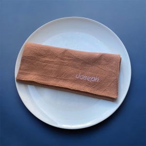 Op de afbeelding: Een gevouwen, roestkleurig linnen servet met de naam "Joseph" in wit garen geborduurd. Het servet ligt op een groot, wit keramisch bord. Het bord staat op een donkerblauwe ondergrond.