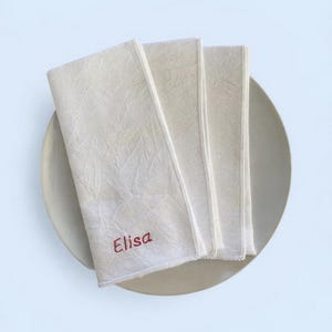 Op de afbeelding: Off-white linnen servetten, opgevouwen en gestapeld op een neutraal bord. Het woord "Elisa" is in het rood op de bovenste servet geborduurd. De servetten hebben een textuur.