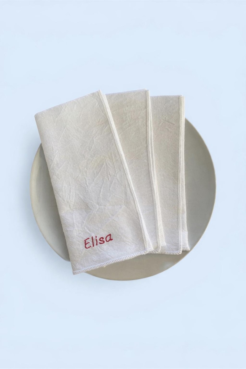 Op de afbeelding: Off-white linnen servetten, opgevouwen en gestapeld op een neutraal bord. Het woord "Elisa" is in het rood op de bovenste servet geborduurd. De servetten hebben een textuur.