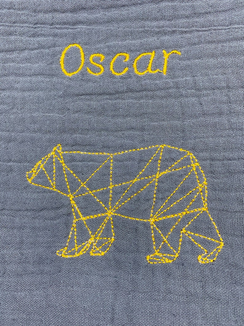May include: A gray fabric baby blanket with the name "Oscar" embroidered in yellow thread. A geometric bear design is also embroidered in yellow thread.