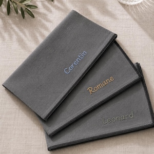 Op de afbeelding: Drie grijze stoffen servetten met geborduurde namen: "Corentin" in blauw, "Romane" in goud en "Leonard" in grijs. Elk servet heeft een donkergrijze rand. De servetten zijn netjes opgevouwen en op een licht oppervlak geplaatst.