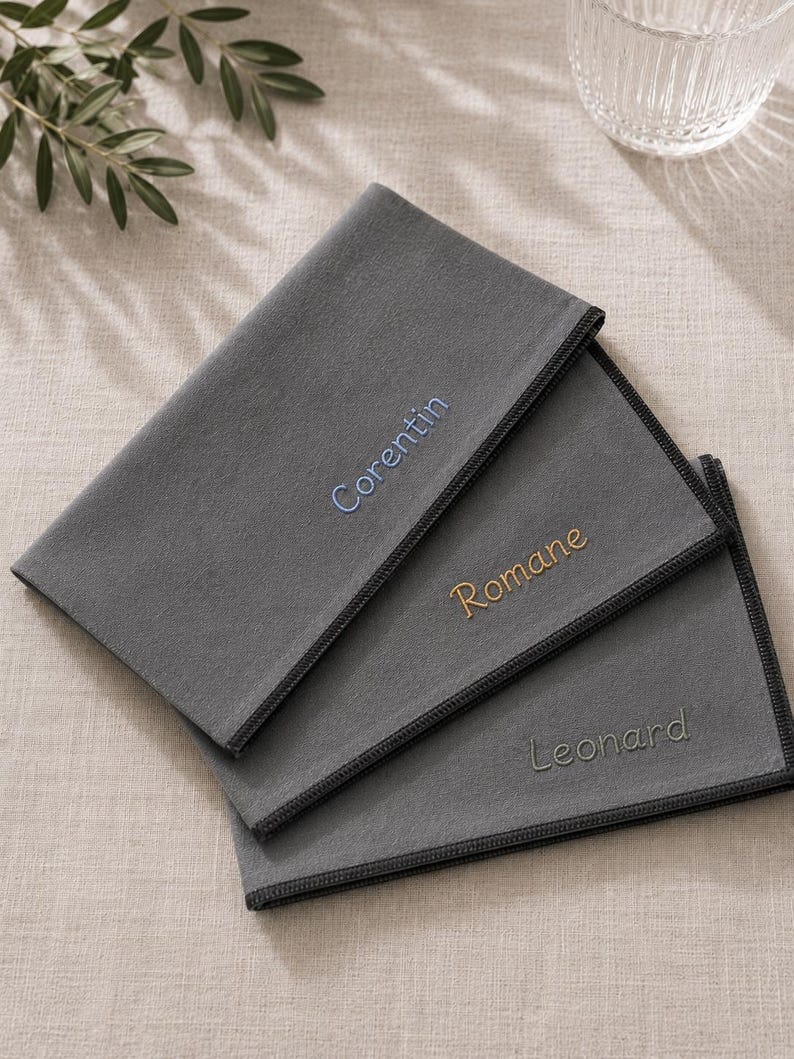 Op de afbeelding: Drie grijze stoffen servetten met geborduurde namen: "Corentin" in blauw, "Romane" in goud en "Leonard" in grijs. Elk servet heeft een donkergrijze rand. De servetten zijn netjes opgevouwen en op een licht oppervlak geplaatst.