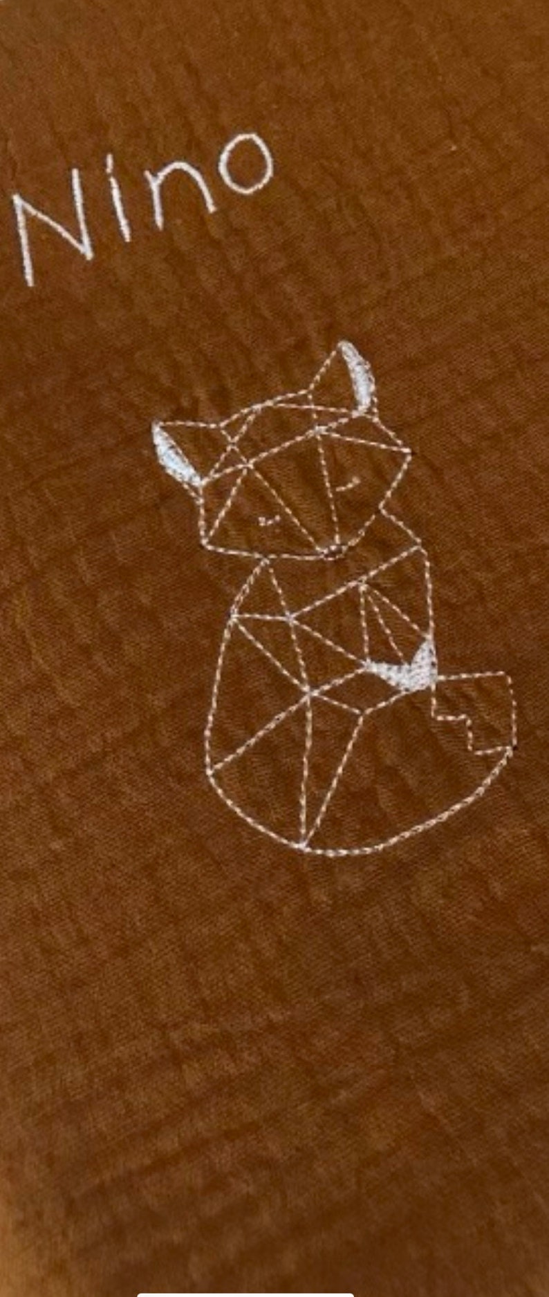 May include: A brown fabric baby blanket with white embroidery of a geometric fox and the word "Nino".