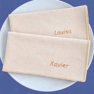 Op de afbeelding: Twee beige gepersonaliseerde servetten met goud geborduurde namen "Louisa" en "Xavier" liggen op een wit bord. De servetten hebben een gouden gestikte rand, tegen een blauwe achtergrond.