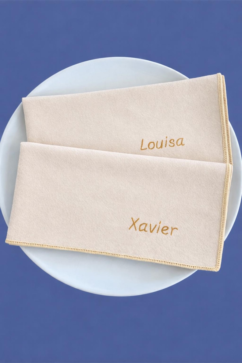 Op de afbeelding: Twee beige gepersonaliseerde servetten met goud geborduurde namen "Louisa" en "Xavier" liggen op een wit bord. De servetten hebben een gouden gestikte rand, tegen een blauwe achtergrond.