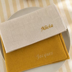 May include: Two personalized linen napkins, one cream and one mustard yellow, embroidered with the names "Alicia" and "Jacques" in gold thread. The napkins are placed on a white plate, set against a striped yellow and white tablecloth.