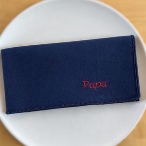 Op de afbeelding: Een marineblauwe rechthoekige doek met het woord "Papa" in het rood geborduurd. De doek is gevouwen en op een wit bord geplaatst. De randen van de doek zijn netjes gestikt.