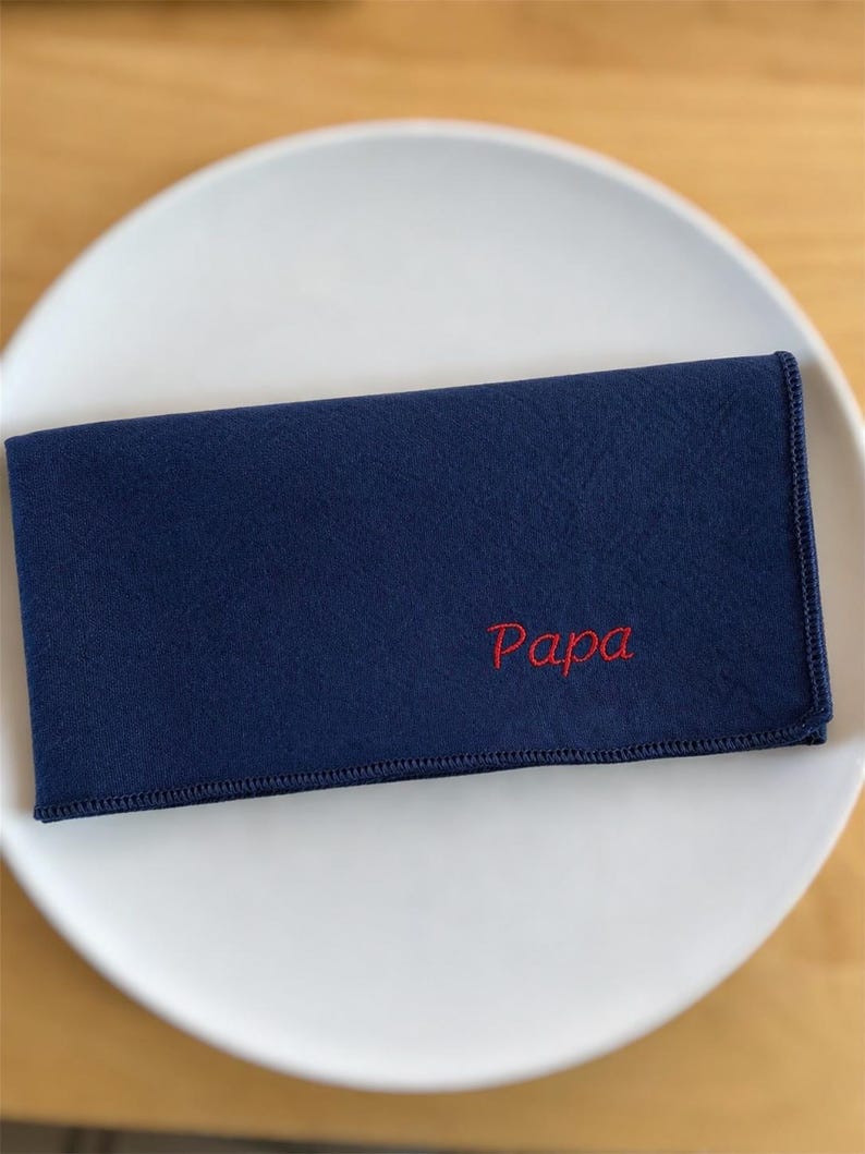 Op de afbeelding: Een marineblauwe rechthoekige doek met het woord "Papa" in het rood geborduurd. De doek is gevouwen en op een wit bord geplaatst. De randen van de doek zijn netjes gestikt.
