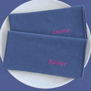 Könnte beinhalten: Zwei blaue Stoffservietten mit den gestickten Namen "Louisa" und "Xavier" in Pink liegen auf einem weißen Teller. Die rechteckigen Servietten haben einen genähten Rand, vor blauem Hintergrund.