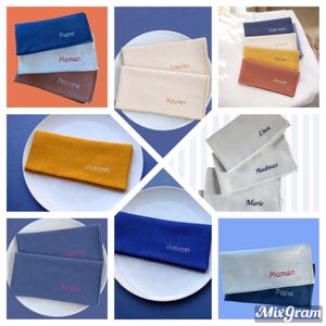 Op de afbeelding: Een collage van gepersonaliseerde stoffen servetten in verschillende kleuren, waaronder blauw, beige en mosterdgeel. Elke servet is geborduurd met een naam of woord in een contrasterende kleur. De servetten zijn gerangschikt op borden.